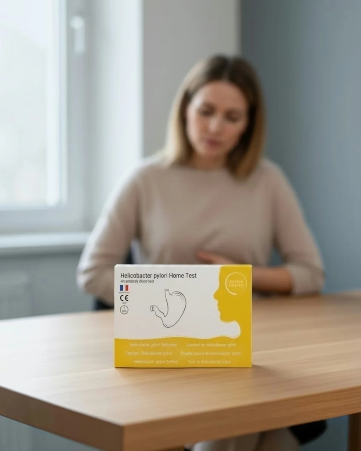 Autotest de Helicobacter pylori Patris Health posé sur une table, avec en arrière-plan une femme souffrant de maux d’estomac